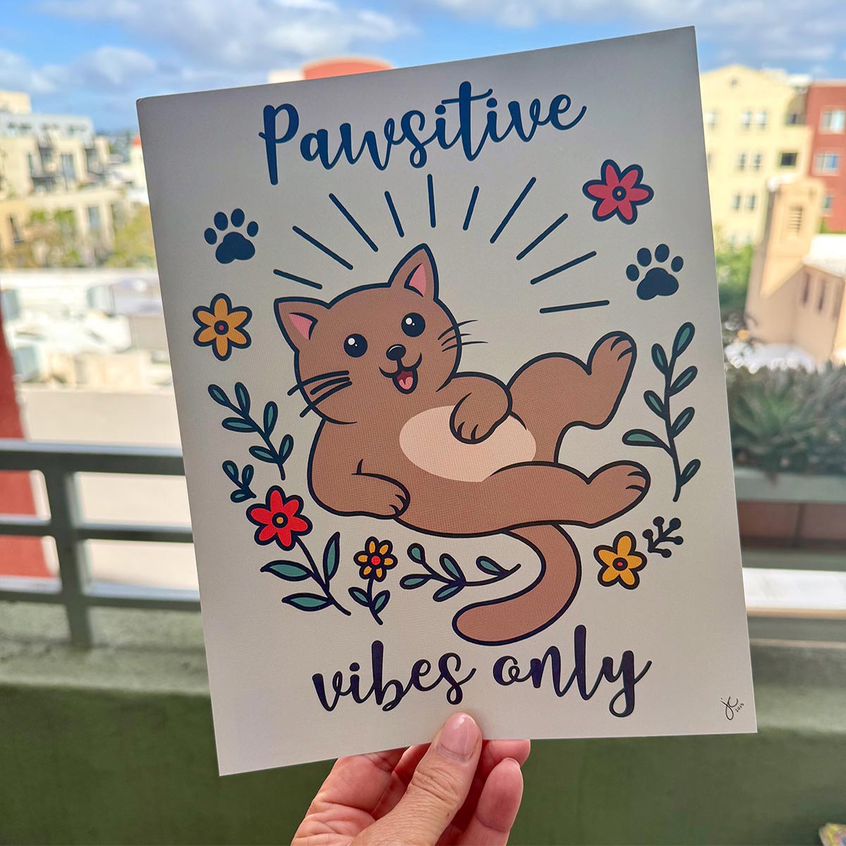 Person holding art print outdoors with city buildings in the background, the Pawsitive Vibes Only art print features a happy cartoon brown cat surrounded in a circle of red and yellow flowers and black paw prints with words Pawsitive on top and vibes on bottom print is on a light muted green background
