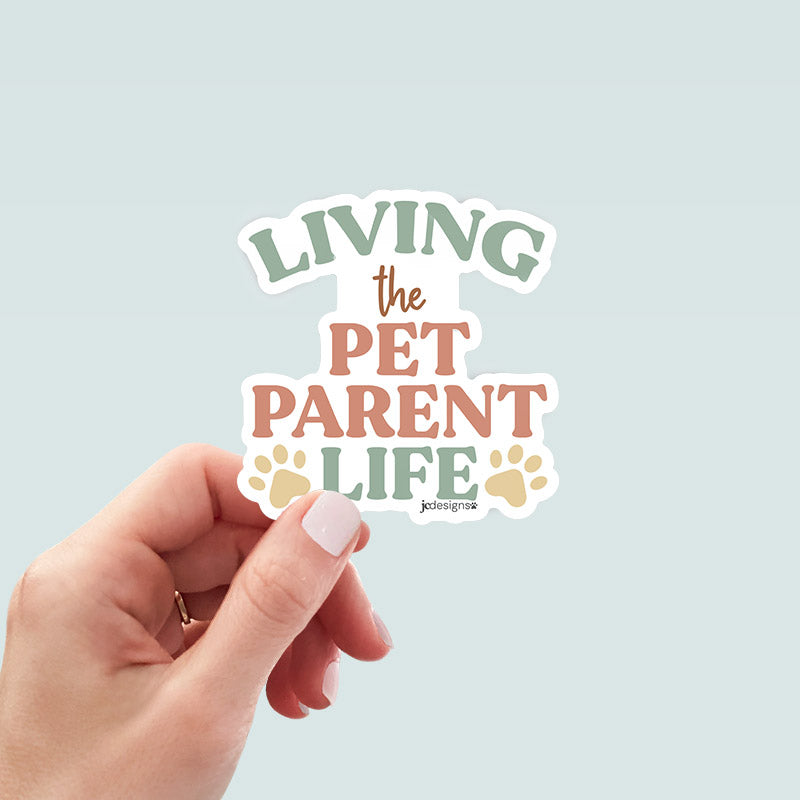 hand holding living the pet parent life sticker with modern typography and paw print accents in earthy pastel colors perfect for cat dad, dog mom