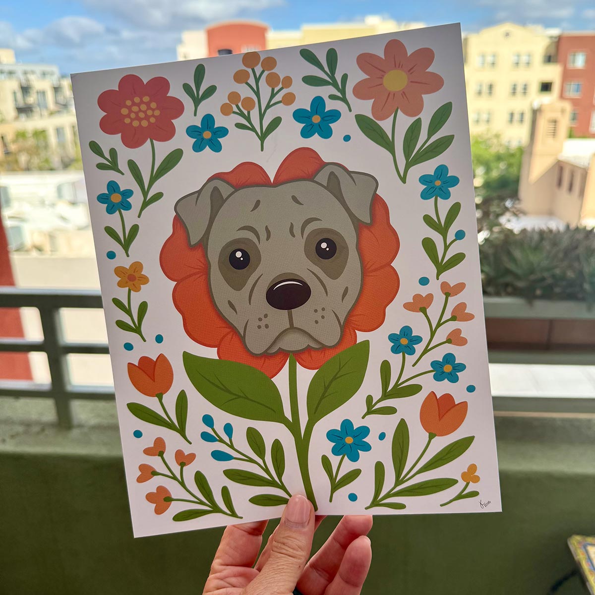 hand holding art print outdoors with colorful city buildings in background, the Pit Bull Flower art print features a gray Pit Bull's face framed by an orange flower surrounded by orange and teal flowers and green stems leaves on a white background