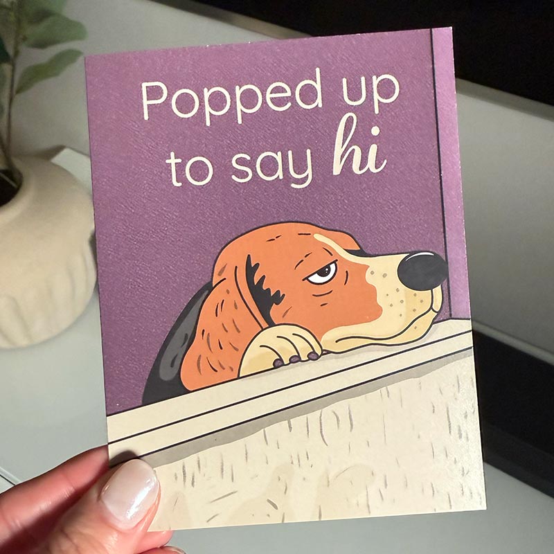 hand holding Illustrated greeting card of a dog popping up over a wall with the text “Popped up to say hi”