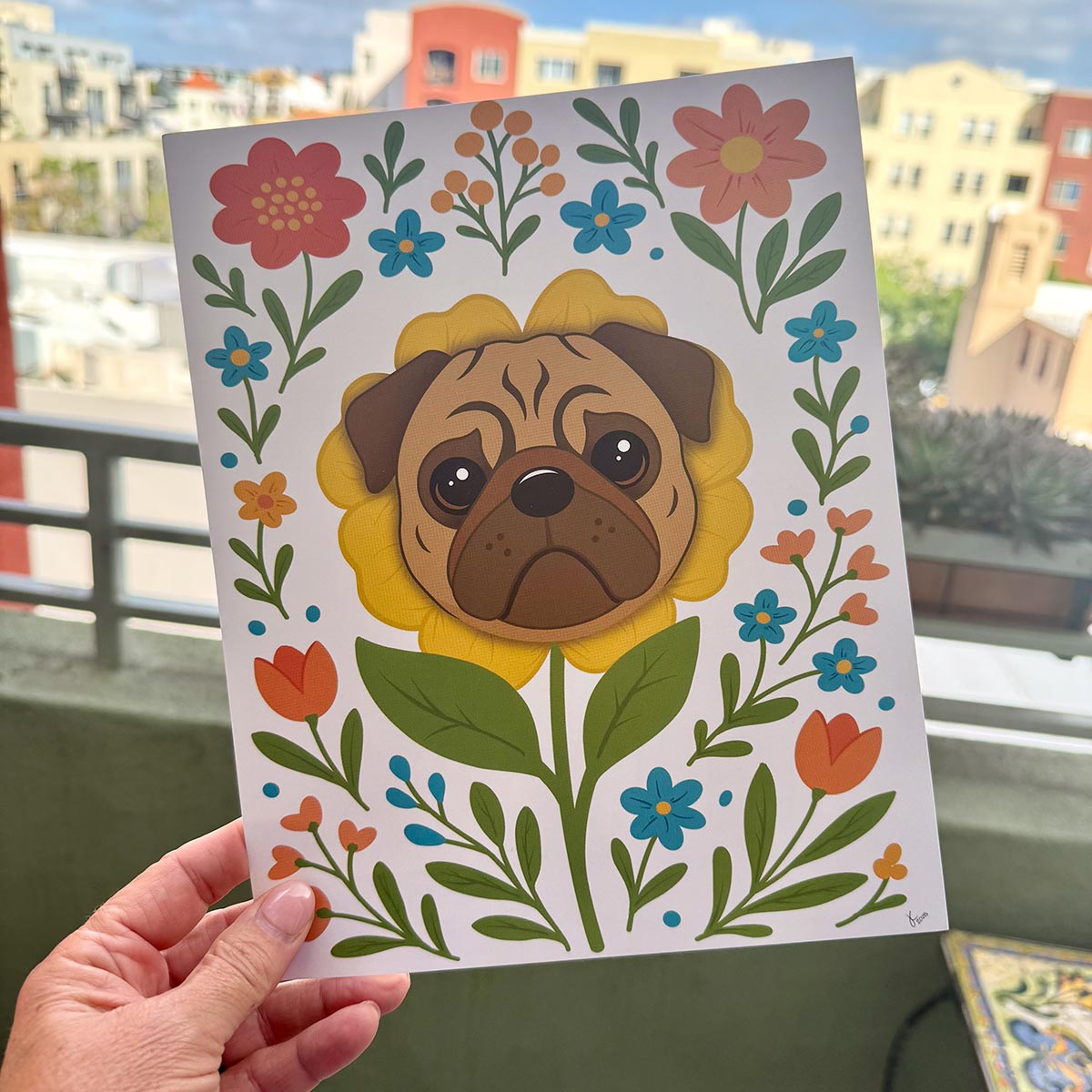 hand holding Pug flower art print outdoors with colorful city buildings in background, art print features a brown pug's face framed by a yellow flower and surrounded by colorful orange yellow and teal blooms with green leaves and stems on a white background