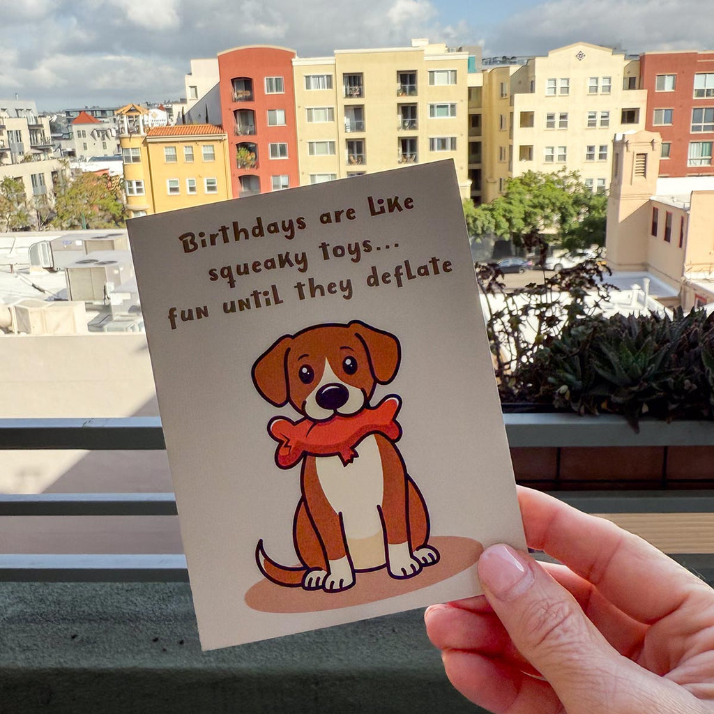 hand holding card outdoors with colorful city buildings in background, the funny Squeaky Toys birthday card features a cute brown and white dog sitting downwith a deflated red squeaky toy with chew tear in its mouth, text on top of card reads "Birthdays are like squeaky toys...fun until they deflate", the card has a light muted brown background