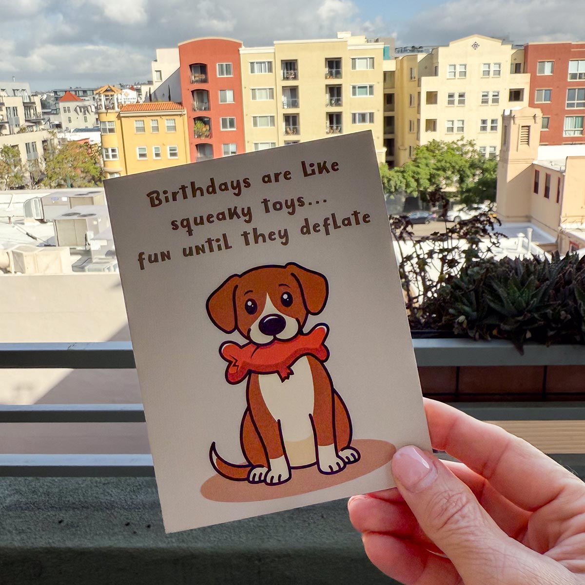 hand holding card outdoors with colorful city buildings in background, the funny Squeaky Toys birthday card features a cute brown and white dog sitting downwith a deflated red squeaky toy with chew tear in its mouth, text on top of card reads "Birthdays are like squeaky toys...fun until they deflate", the card has a light muted brown background