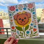 hand holding art print outdoors with colorful city buildings in the background, the Terrier Flower art print features a brown terrier face framed by an yellow flower surrounded by orange and teal flowers and green stems leaves on a white background