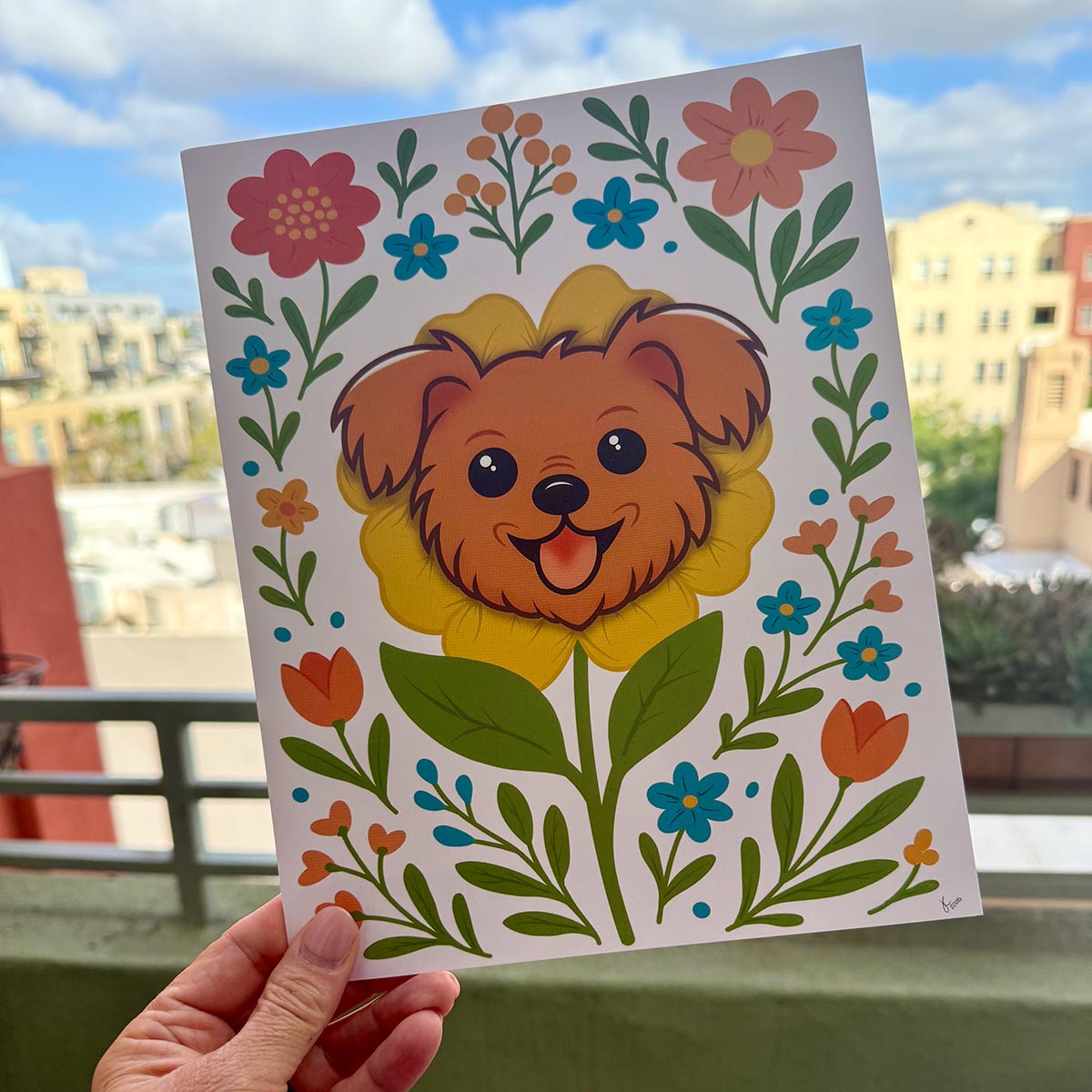 hand holding art print outdoors with colorful city buildings in the background, the Terrier Flower art print features a brown terrier face framed by an yellow flower surrounded by orange and teal flowers and green stems leaves on a white background