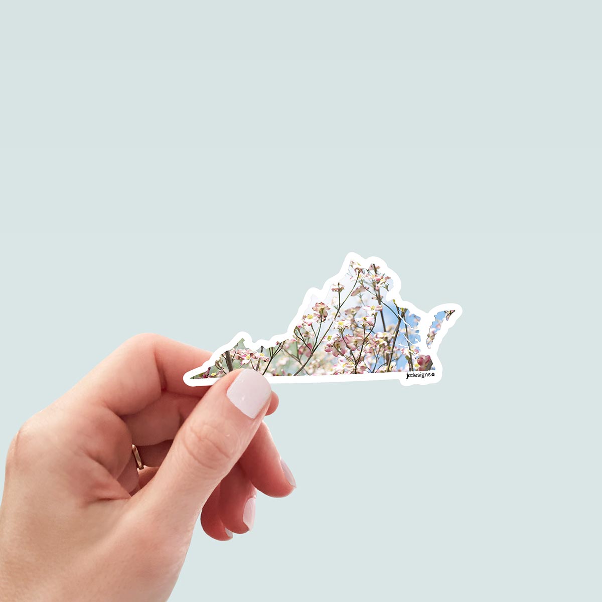 hand holding Virginia state-shaped sticker featuring blooming pink and white dogwood flowers with a light blue sky background, outlined in white die cut
