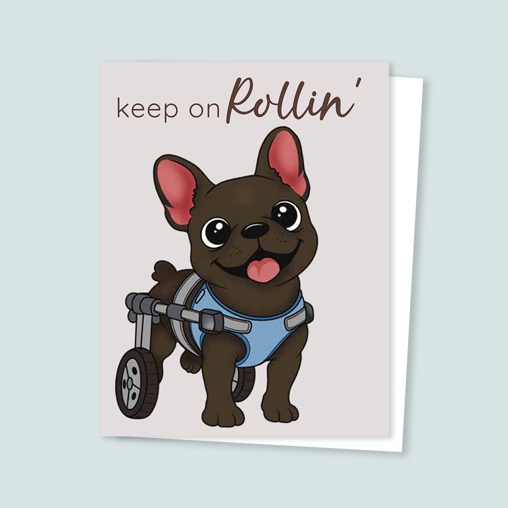 Hand-illustrated greeting card featuring a happy brown cartoon dog wearing a blue mobility wheelchair harness with two back wheels, smiling with tongue out under the words “Keep on Rollin’.” The design conveys encouragement, resilience, and positivity.
