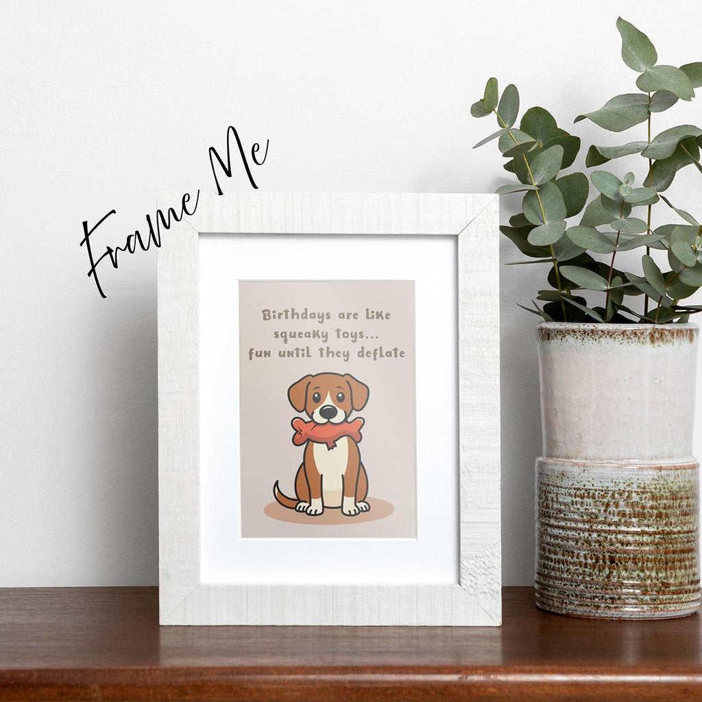 framed Squeaky Toys birthday card featuring a cute brown and white dog sitting downwith a deflated red squeaky toy with chew tear in its mouth, text on top of card reads "Birthdays are like squeaky toys...fun until they deflate", the card has a light muted brown background, frame not included in purchase