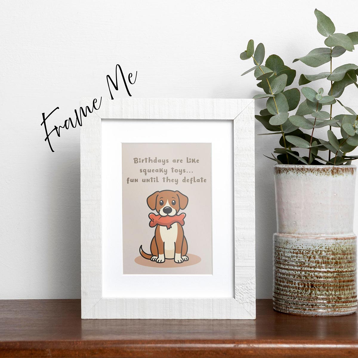 framed Squeaky Toys birthday card featuring a cute brown and white dog sitting downwith a deflated red squeaky toy with chew tear in its mouth, text on top of card reads "Birthdays are like squeaky toys...fun until they deflate", the card has a light muted brown background, frame not included in purchase