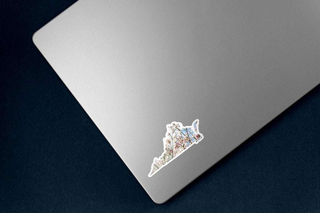 Virginia state-shaped sticker featuring blooming pink and white dogwood flowers with a light blue sky background, outlined in white die cut, sticker on lower left corner of silver laptop